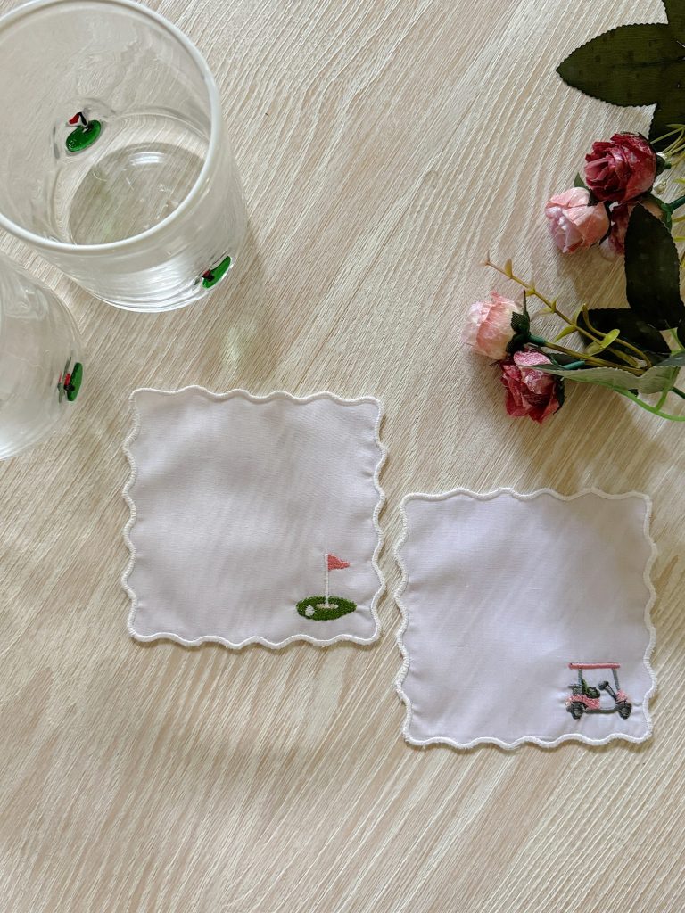 Two clear glasses and two white coasters with embroidered golf-themed designs are on a light wood table next to a bunch of pink artificial flowers.