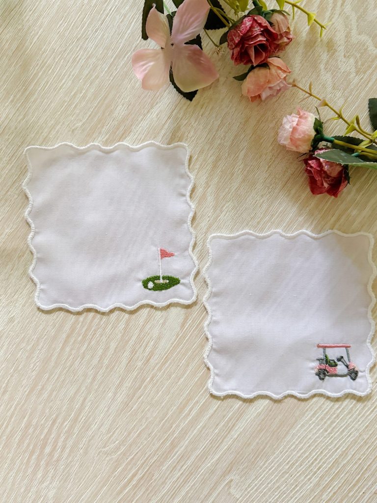 Two white, scallop-edged cloth napkins on a light wood surface; one has a golf flag and green, the other a golf cart. Artificial flowers are arranged in the upper right corner.