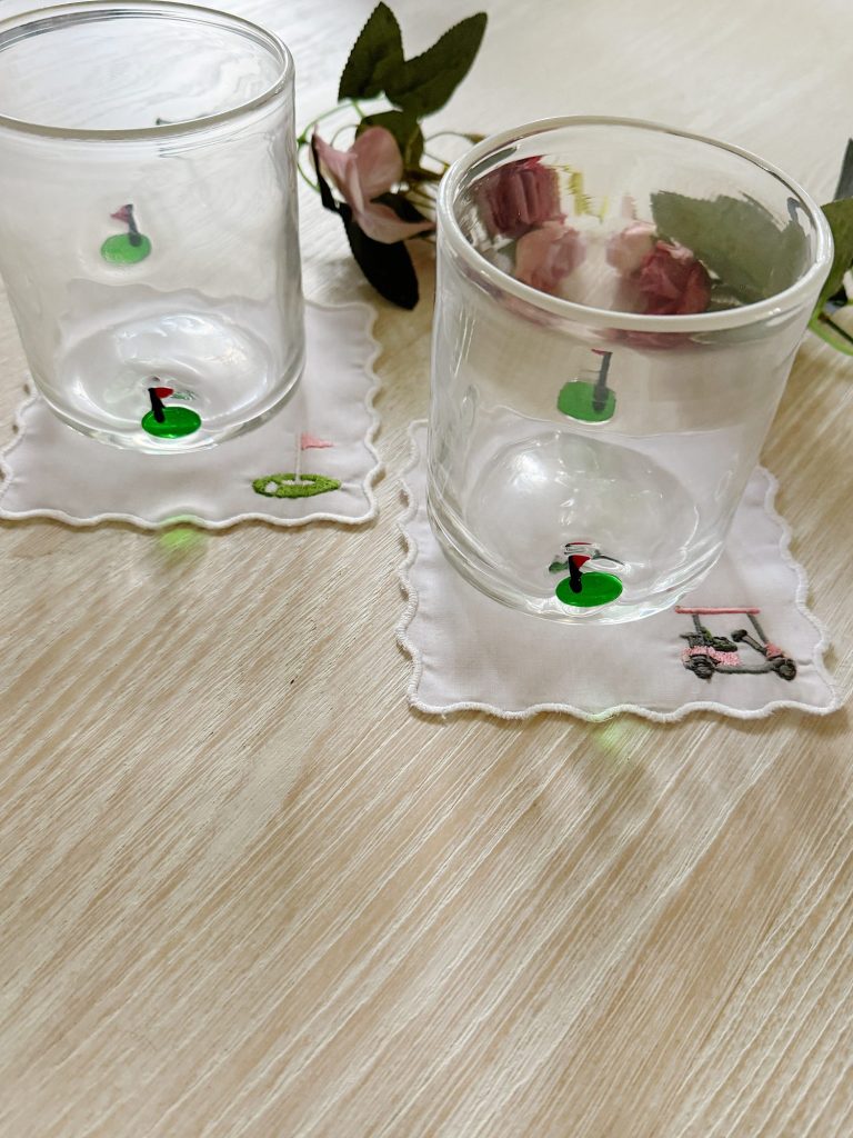 Two empty glass tumblers sit on white embroidered coasters with golf-themed designs, placed on a light wooden surface with artificial flowers nearby.