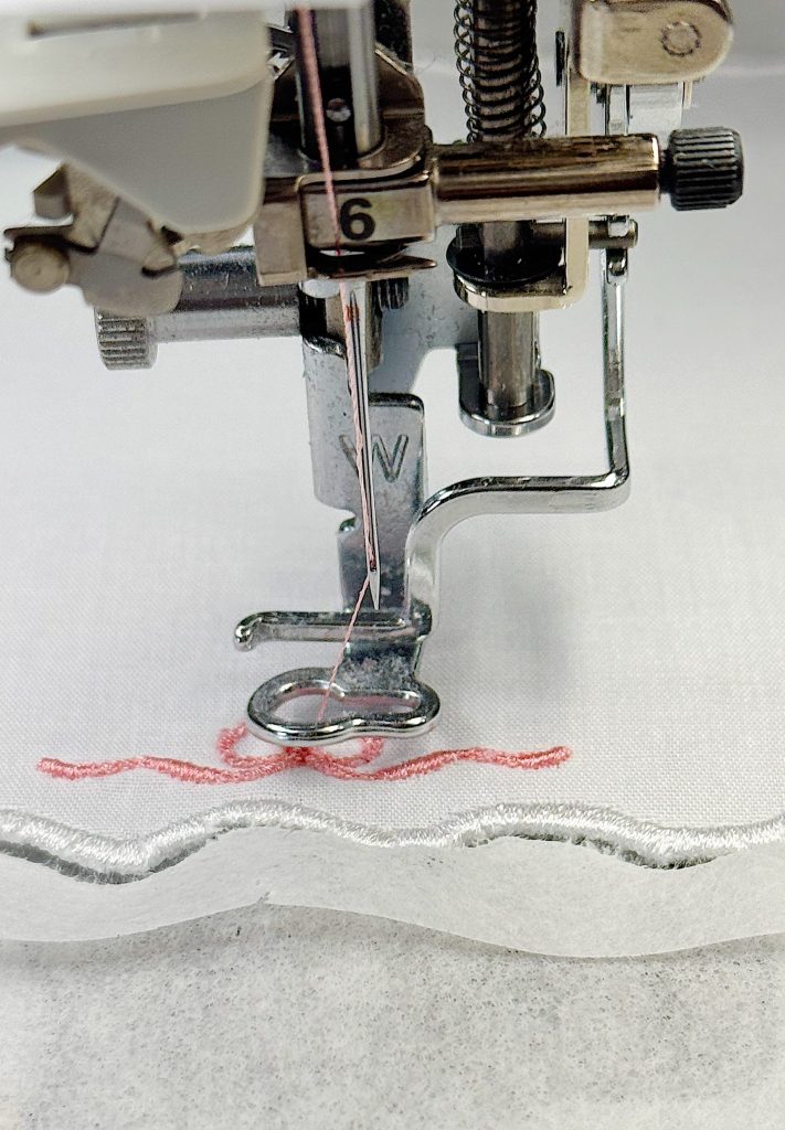 Close-up of a sewing machine needle stitching pink thread onto white fabric with a scalloped edge.