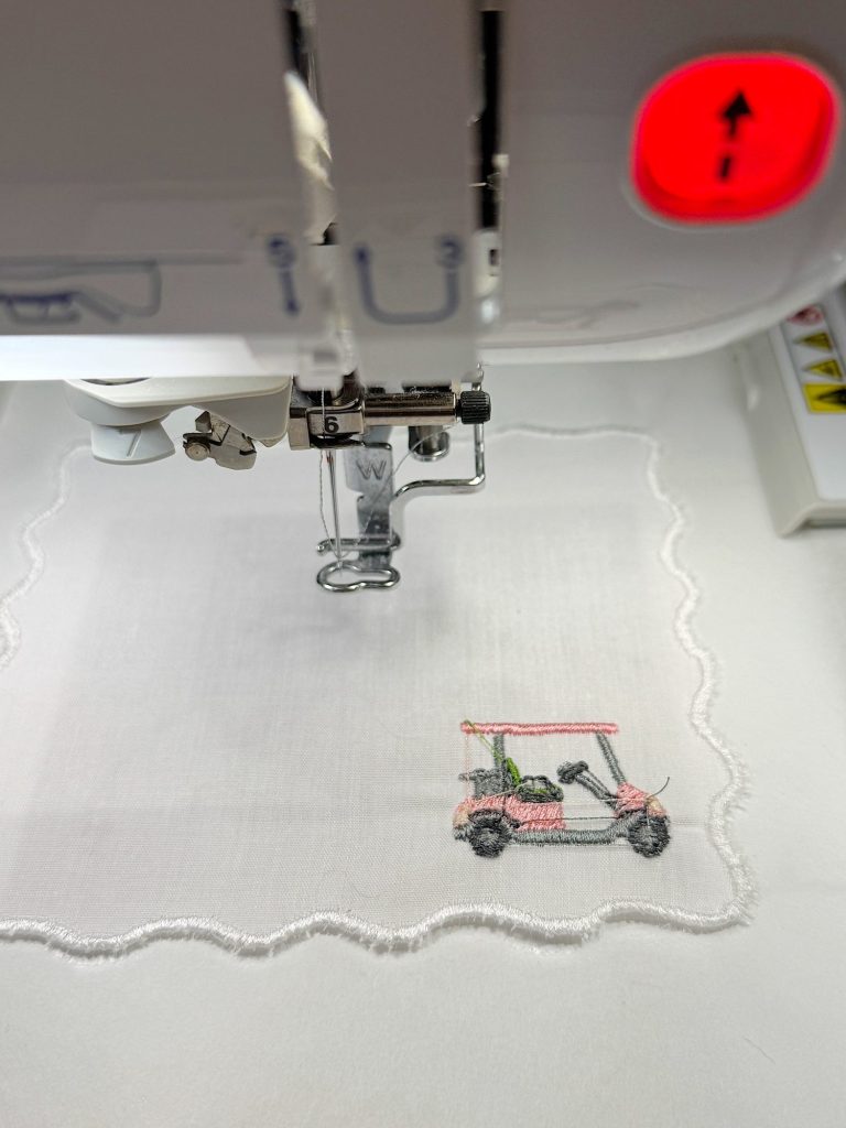 A sewing machine embroidering a design of a golf cart on a white fabric with a scalloped edge.