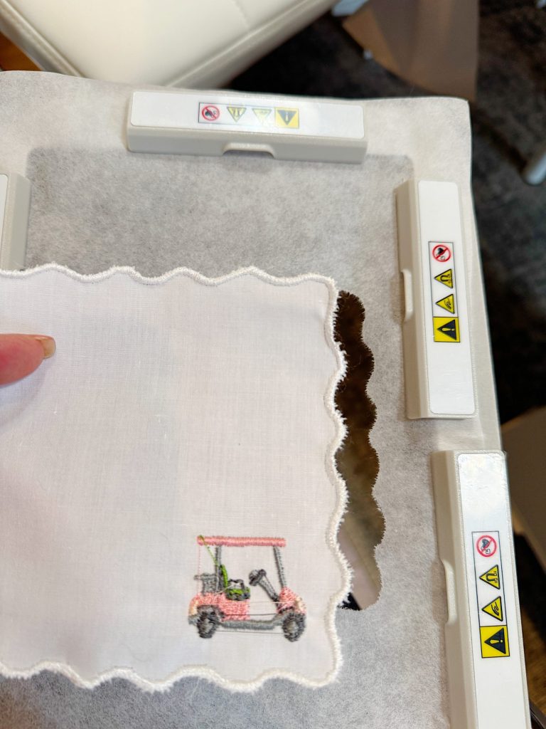 A piece of white fabric with scalloped edges and a small embroidered golf cart is held in an embroidery machine hoop with visible warning labels.