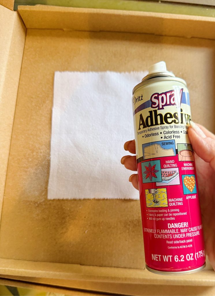 A hand holds a can of spray adhesive over a piece of white fabric placed inside a cardboard box, preparing for application.
