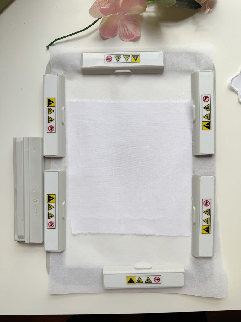 A square piece of white fabric is secured by six white plastic clamps with caution symbols, placed on a white surface with a pink flower in the background.
