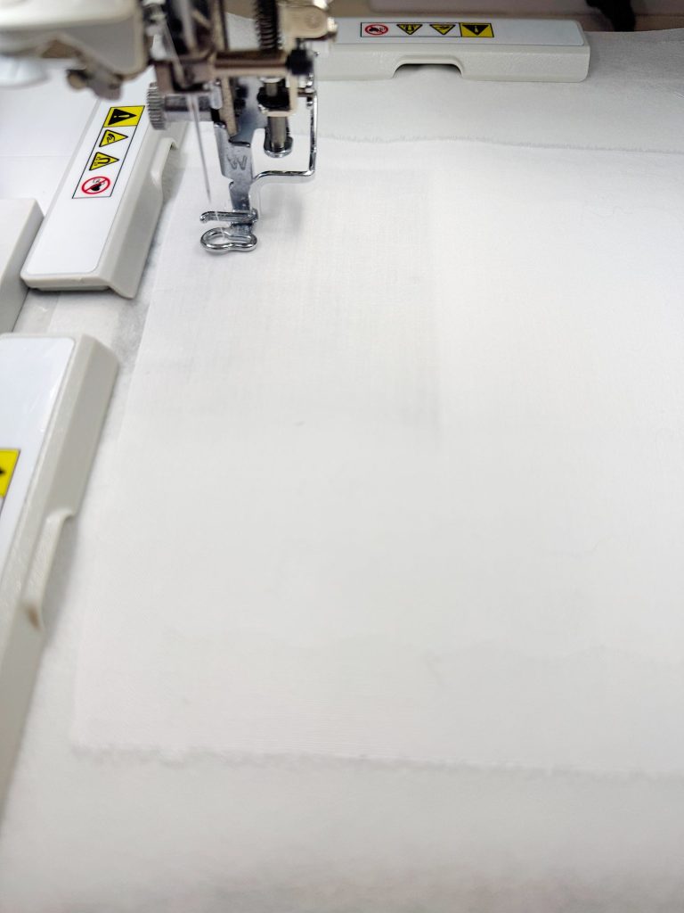Close-up of a sewing machine needle and presser foot stitching white fabric, with safety warning labels visible on machine parts.