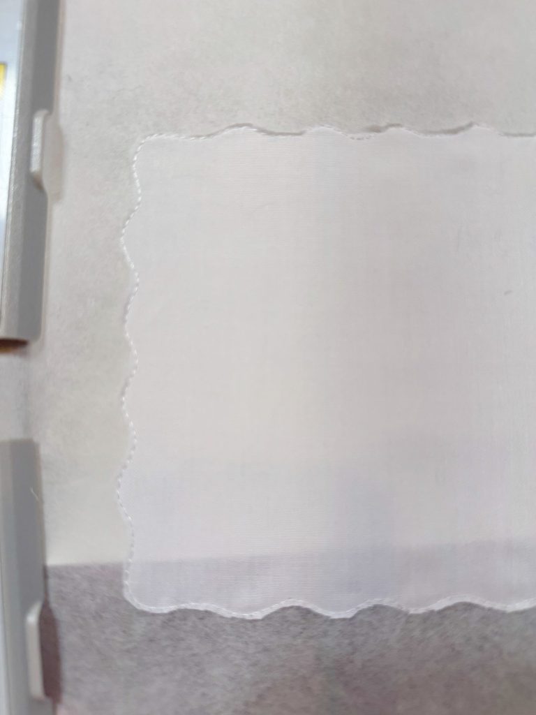 A white fabric swatch with scalloped edges is displayed on a light gray background, partially framed by gray plastic holders on the left side.