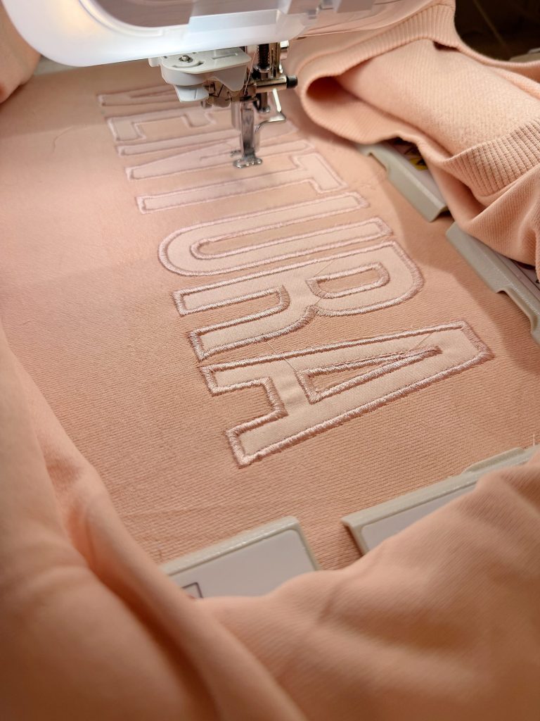 A sewing machine embroiders large outlined letters onto a peach-colored fabric held in place by an embroidery hoop.