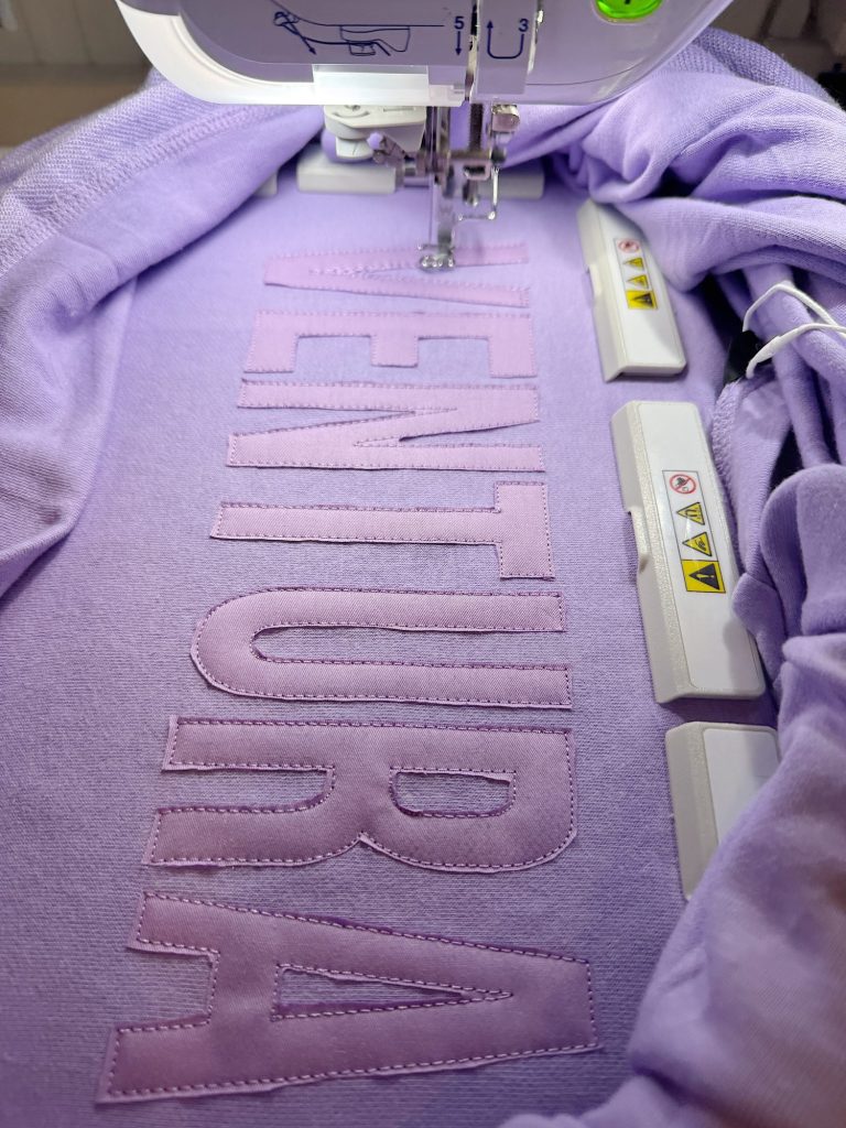 Close-up of an embroidery machine stitching the word "VENTURA" in large letters onto a light purple fabric.