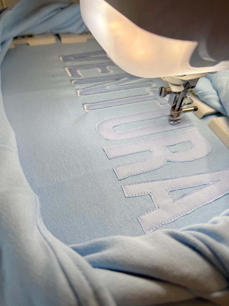 A sewing machine embroiders large, white letters onto a light blue fabric.