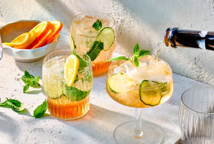 Three cocktails with ice, cucumber, mint, and lemon slices sit on a sunlit table as a beverage is poured from a bottle; a bowl of orange slices is nearby.