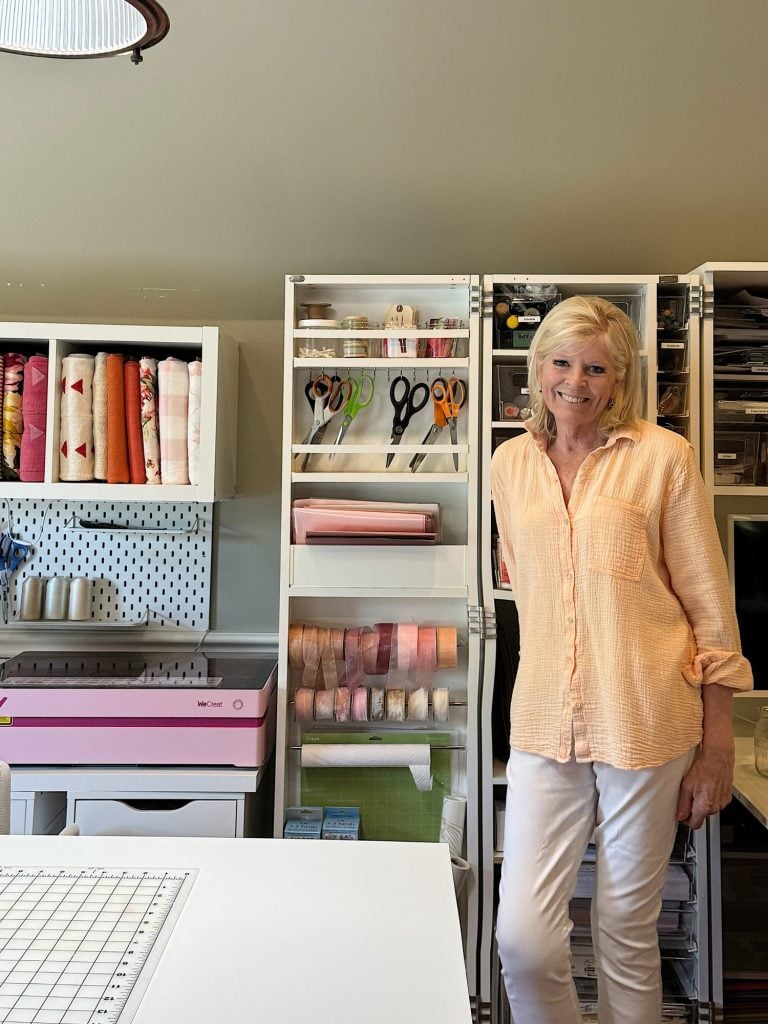 A woman stands in a craft room next to organized shelves holding fabric, scissors, ribbon, and other crafting supplies.