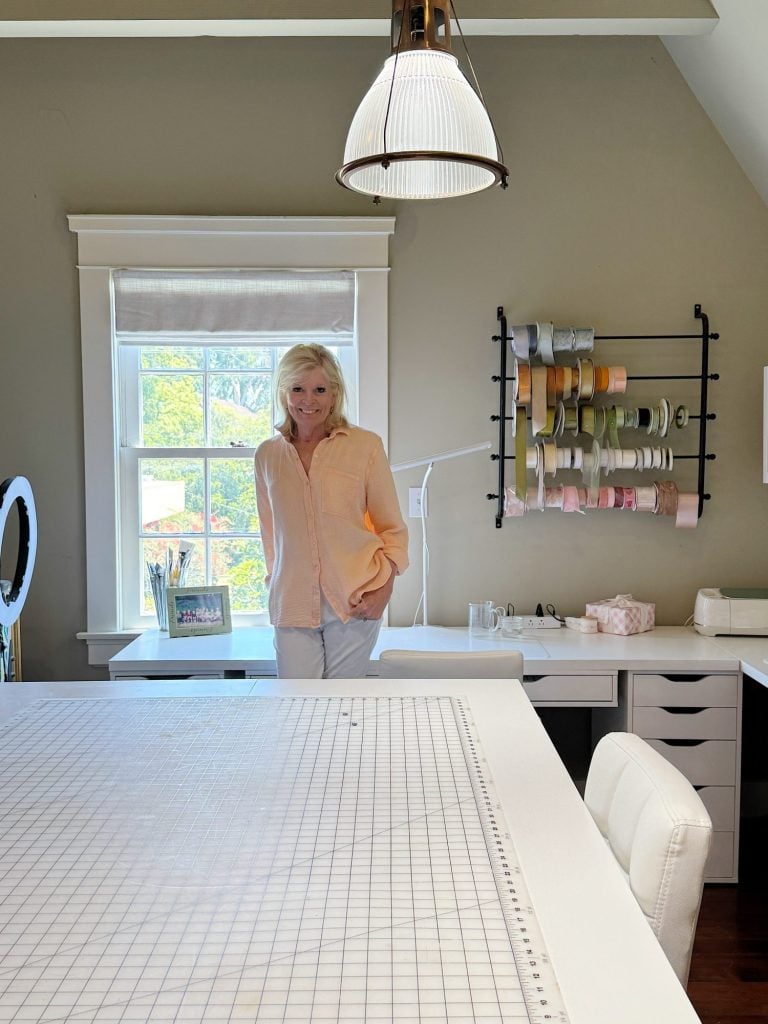 A woman stands in a bright, organized craft room with a large worktable, shelves of ribbon, and a window letting in natural light.