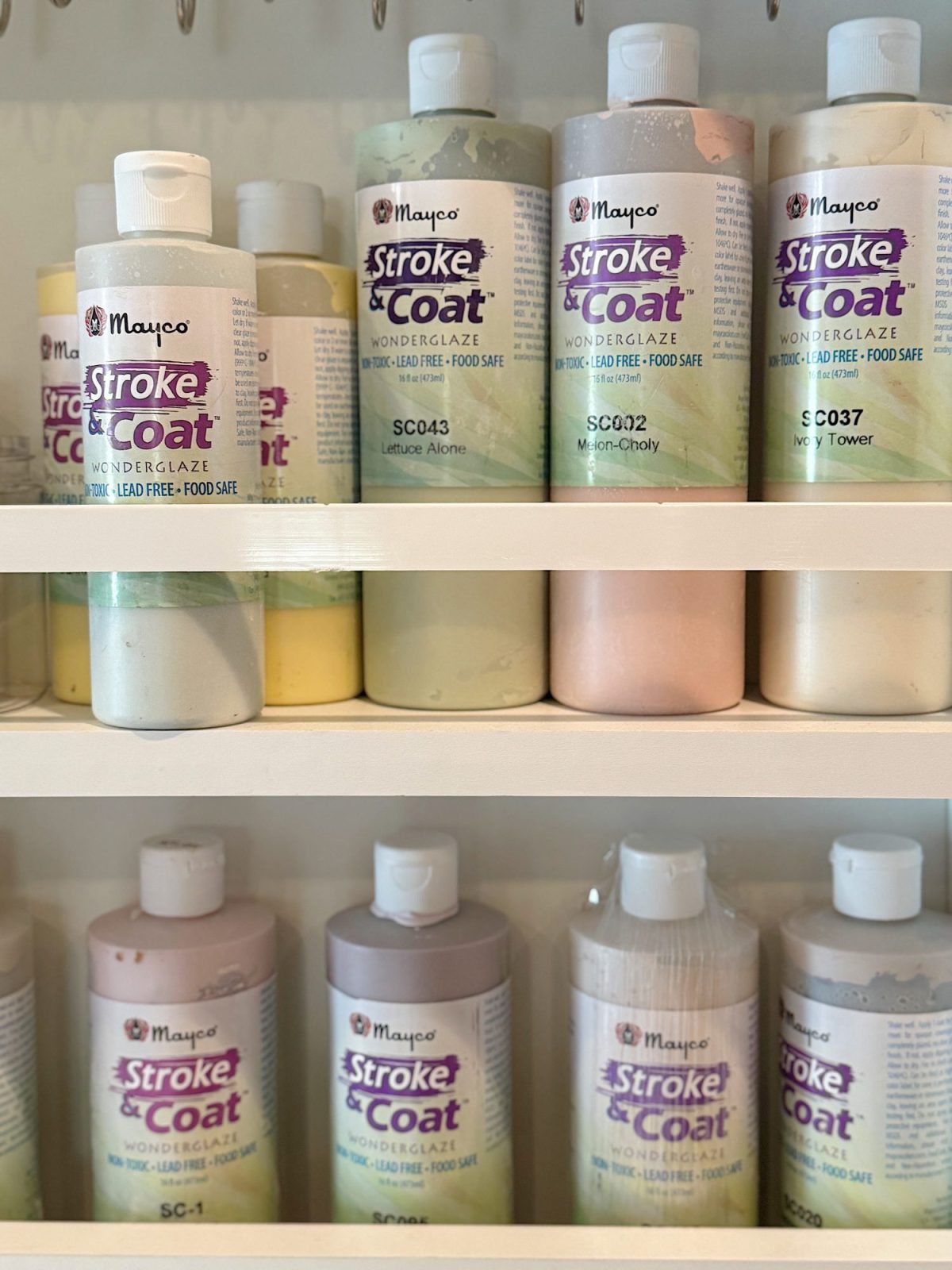 Rows of Mayco Stroke & Coat glaze bottles in various pastel colors arranged neatly on white shelves.