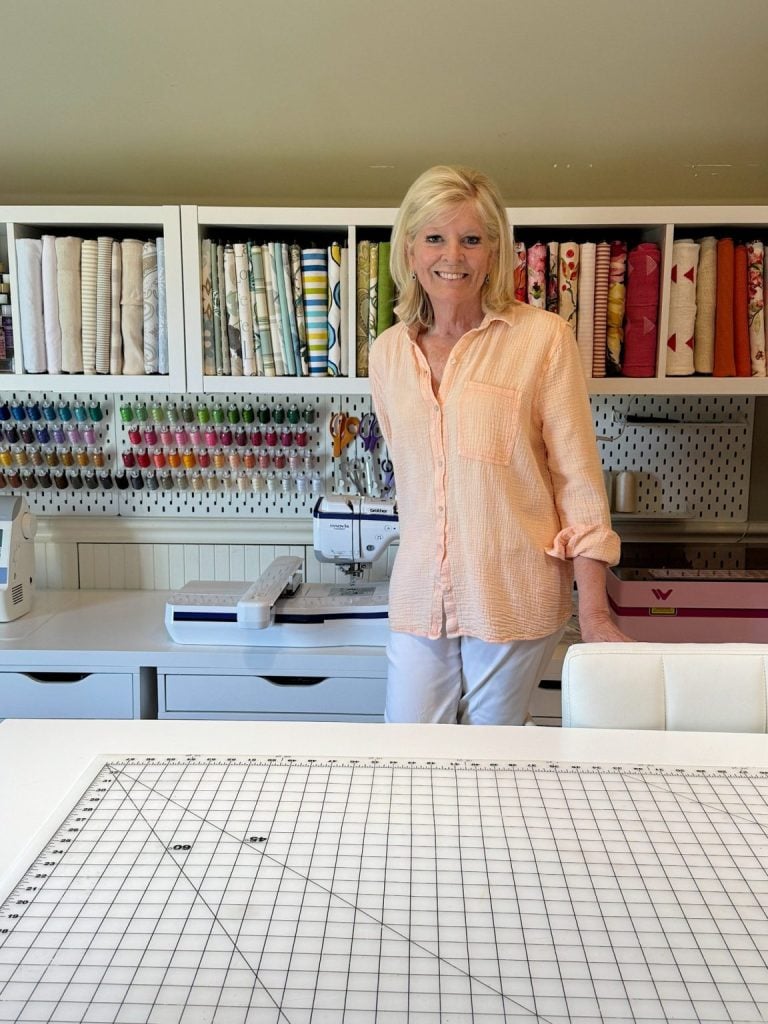 A woman stands in a sewing room with shelves of neatly arranged fabric, rows of thread spools, a sewing machine, and a cutting mat on the table in front of her.