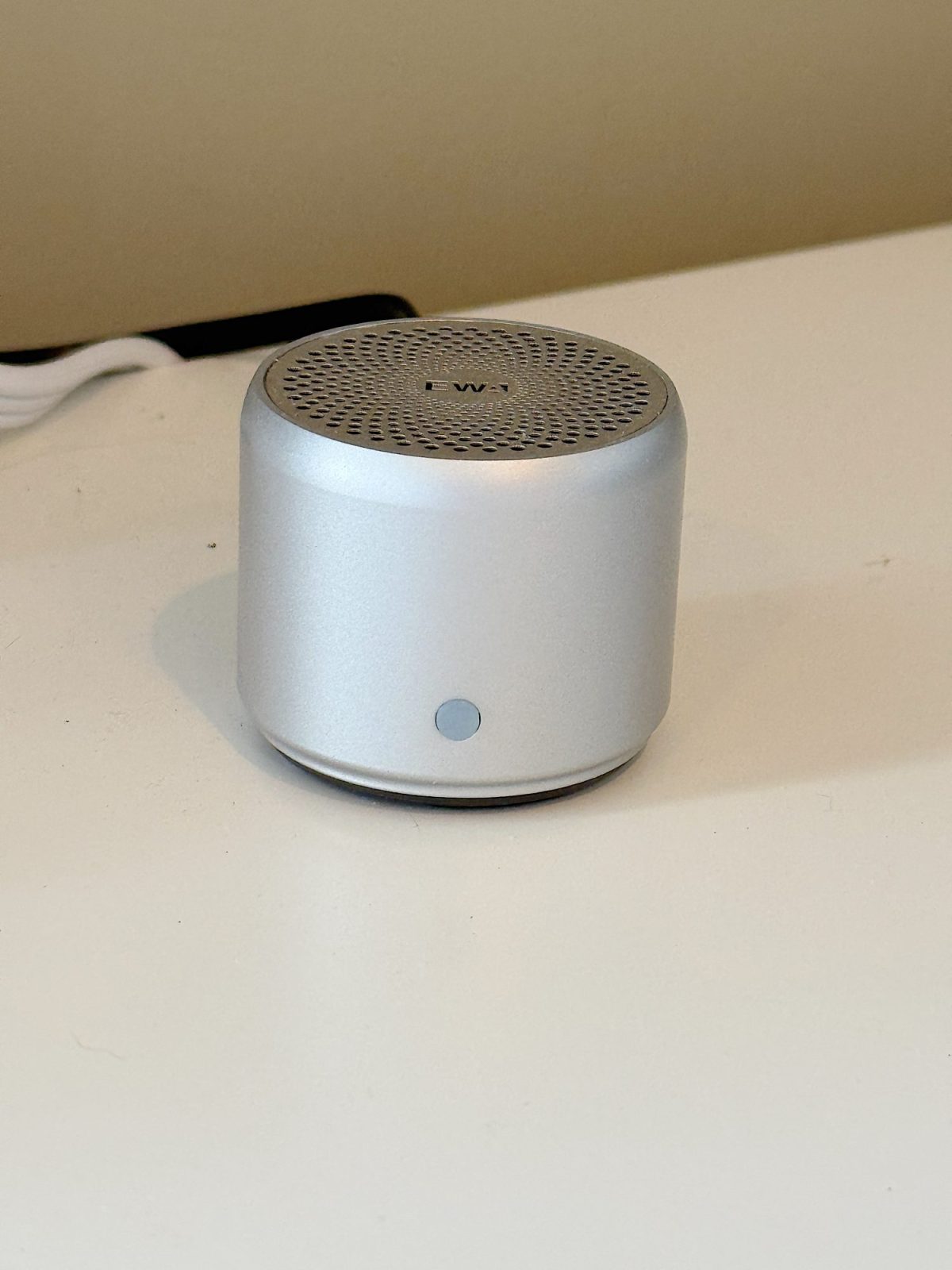 A small, cylindrical silver wireless speaker sits on a white surface near a wall.