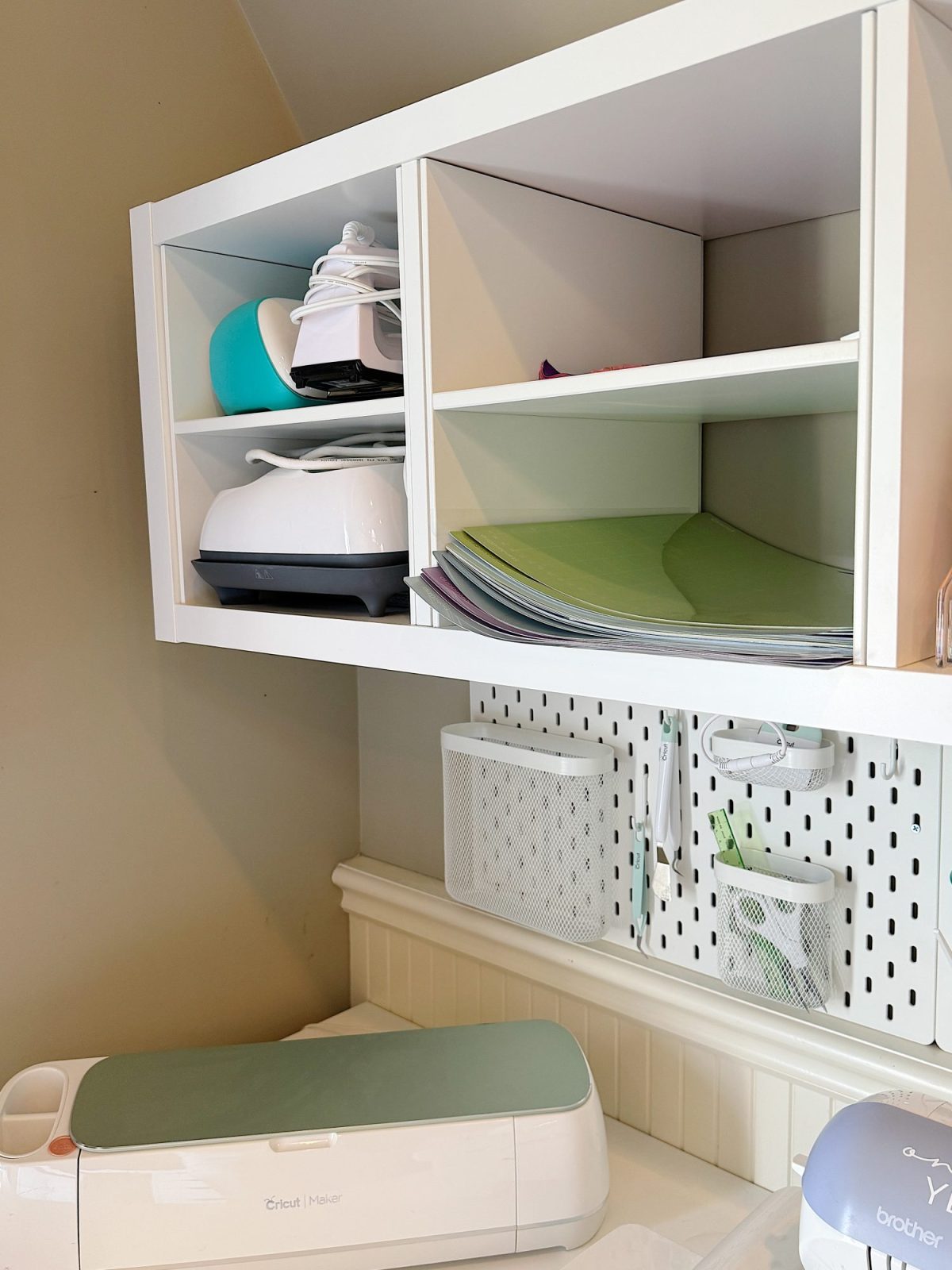 White shelves hold crafting tools, machines, and colored sheets. A pegboard with baskets and a Cricut Maker machine are also visible in the workspace.