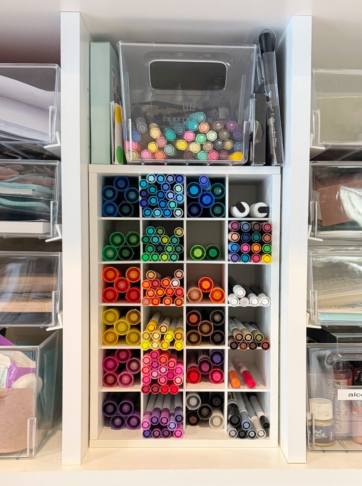 A neatly organized shelf with compartments filled with markers sorted by color, and clear containers holding additional art supplies on either side.