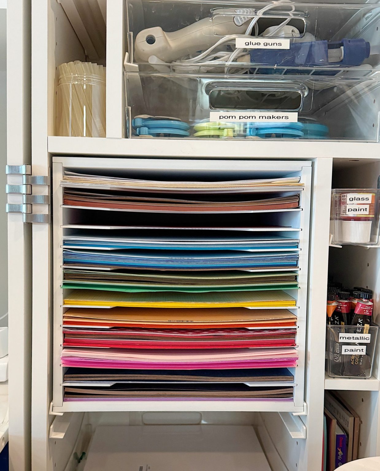 A shelf holds stacked colored paper, organized by shade. Above are trays labeled "glue guns" and "pom pom makers," and beside are labeled containers of glass and metallic paint.