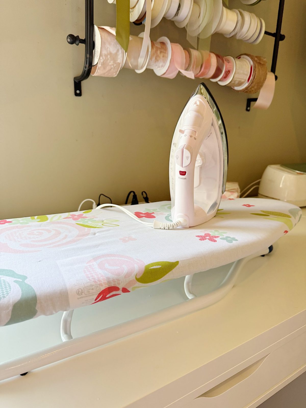 A white iron rests on a small ironing board with a floral cover; rolls of ribbon are stored on a wall rack in the background.