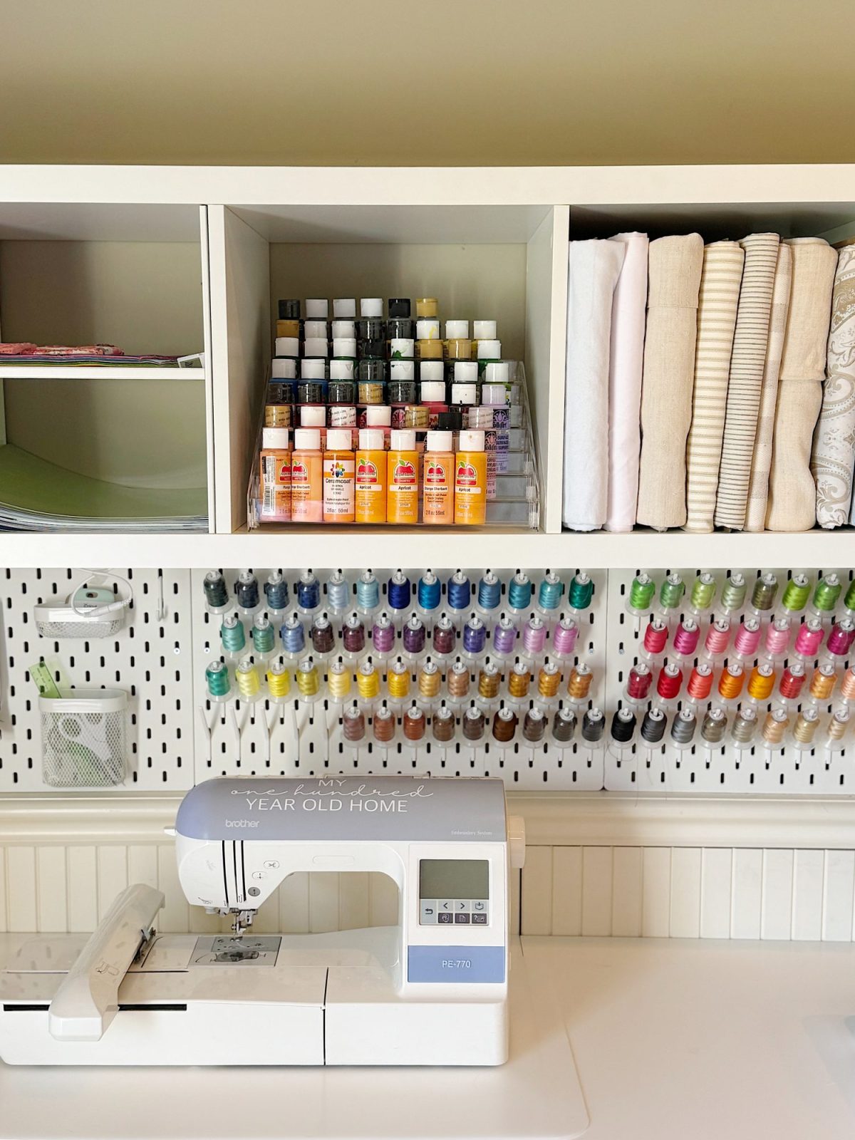 A sewing workstation with a sewing machine, shelves holding fabric and paint bottles, and a pegboard organized with colorful spools of thread.
