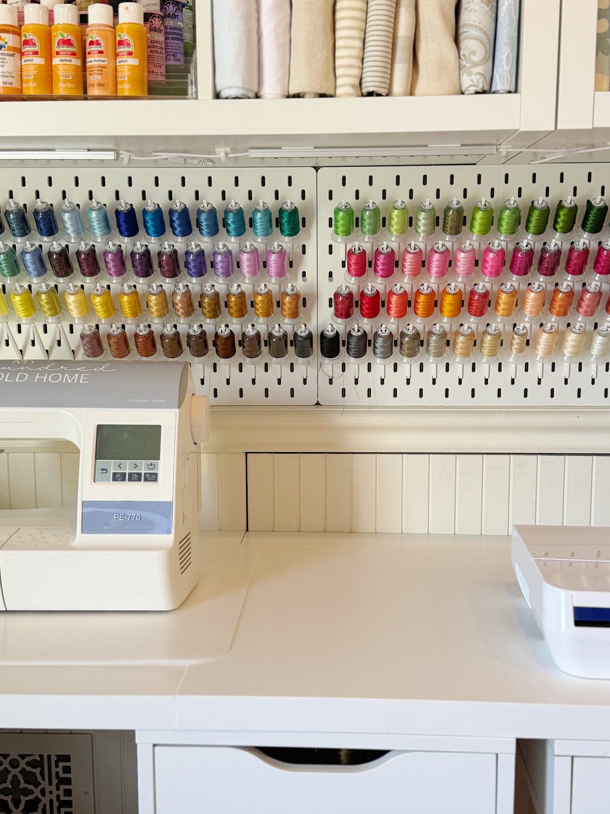 A sewing station with a sewing machine, white desk, and spools of colorful thread organized on a pegboard above the workspace.