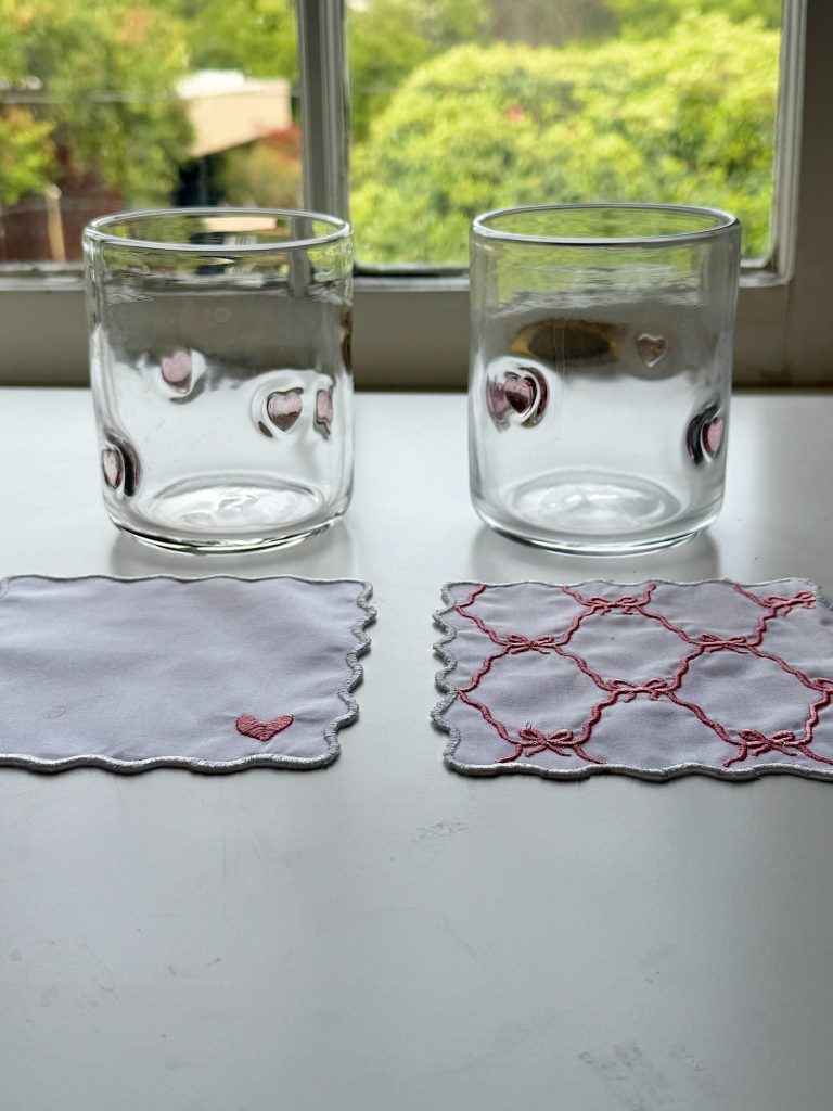 Two clear glasses with heart designs are placed on a white surface in front of a window. Each glass sits on a small, scalloped-edge cloth coaster with pink embroidery.