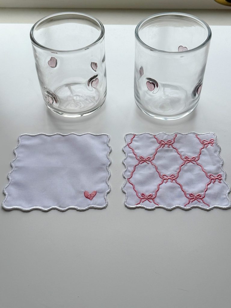 Two clear glasses with pink peach designs are placed above two white fabric coasters, one plain with a pink butterfly, and one with a pink bow pattern.