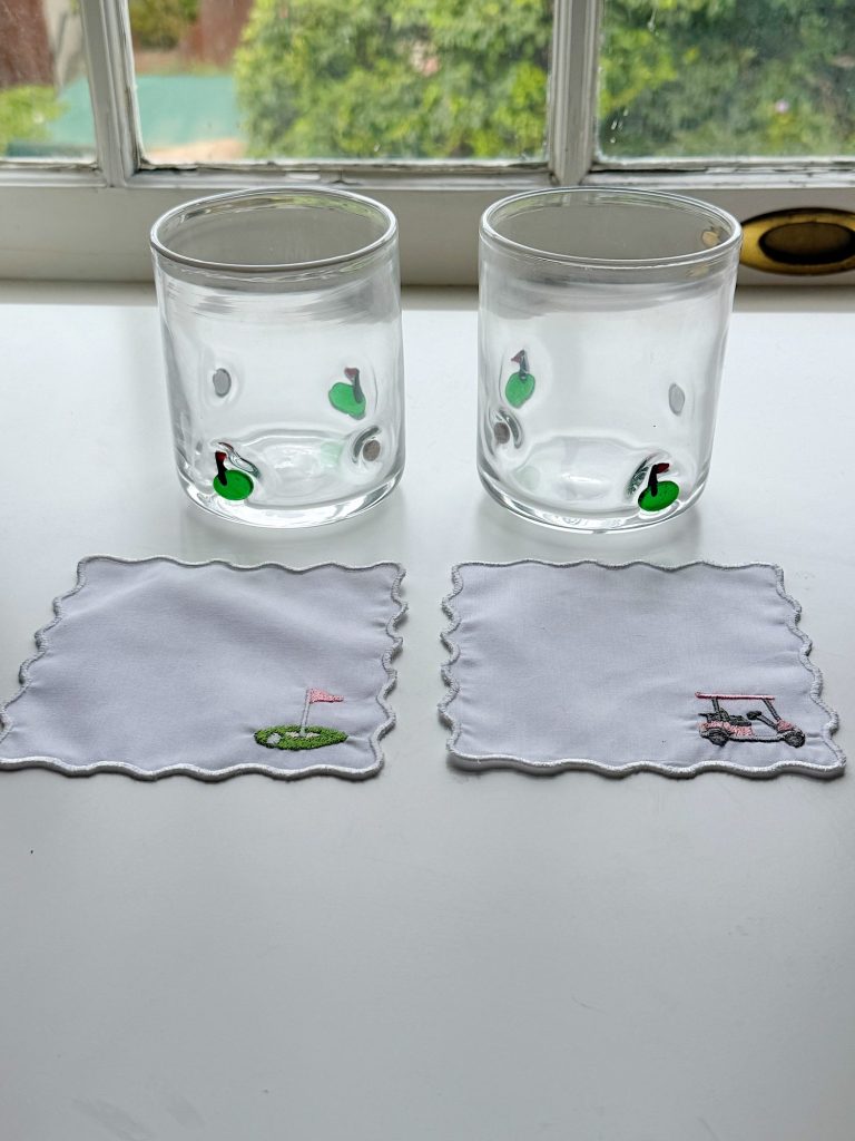 Two clear glasses with green golf-themed designs and two white cloth coasters, one with a golf green and flag, the other with a golf cart, on a white surface by a window.