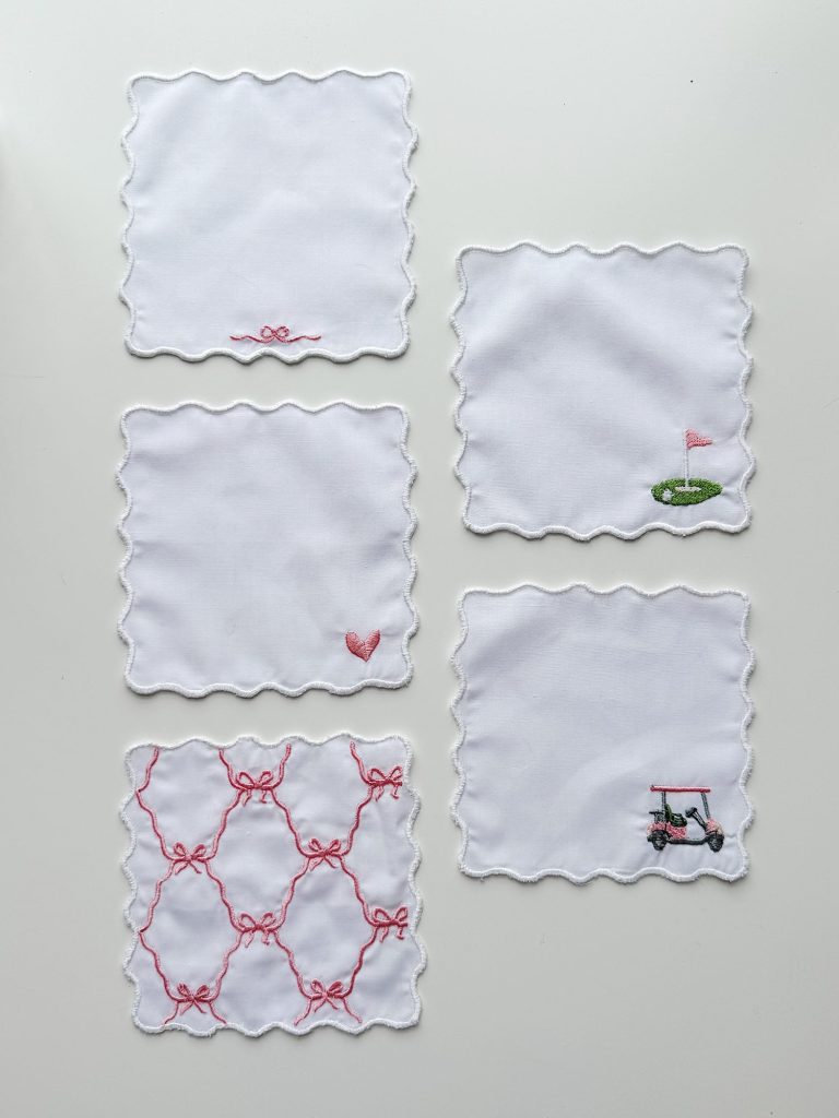 Five white fabric coasters with scalloped edges; three have minimal embroidery, including a golf cart, golf green, and pink designs.