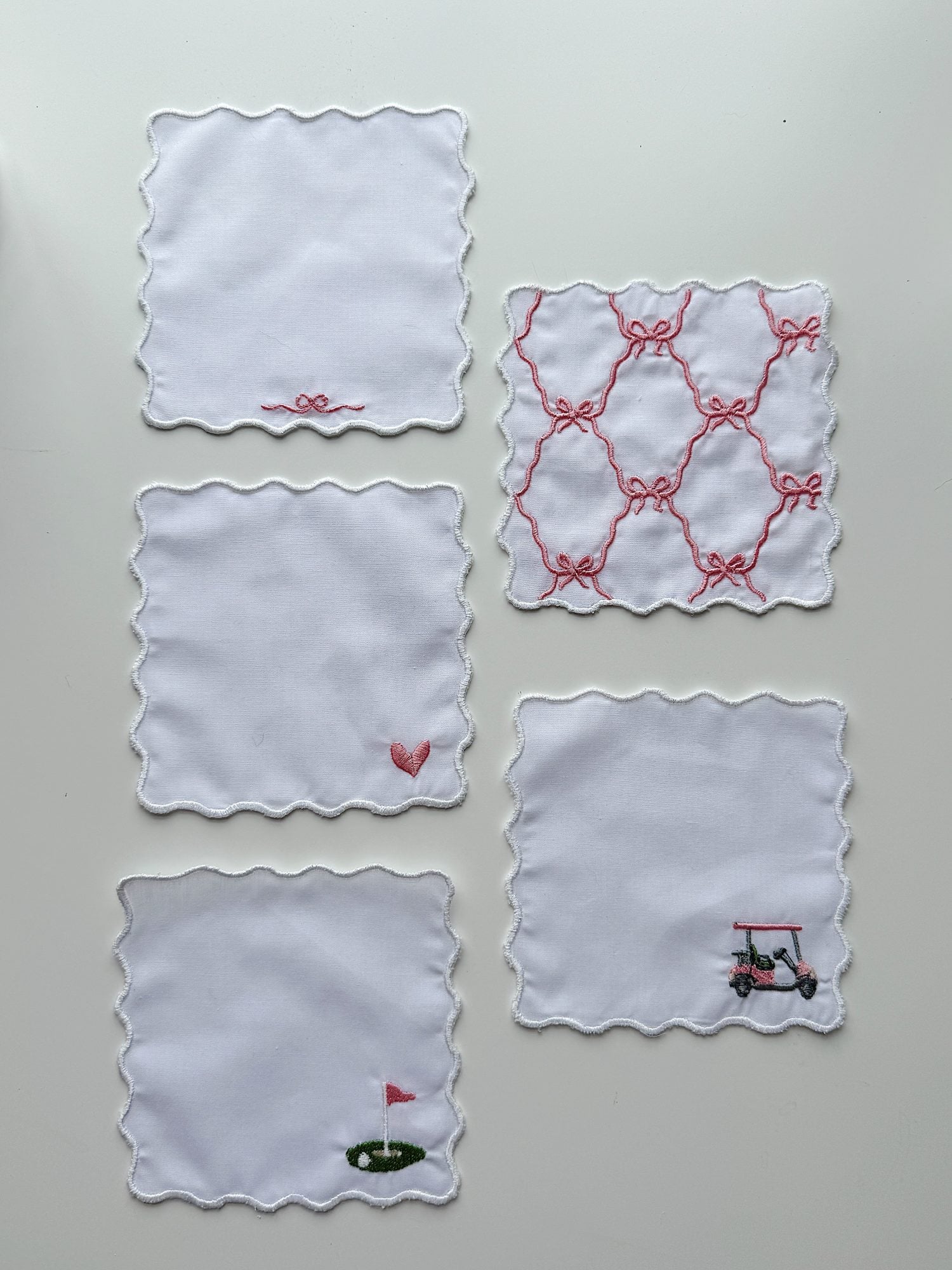 Five white scalloped-edge cloth napkins are arranged on a flat surface, each with a unique pink or red embroidered design, including a heart, golf cart, flag, and geometric pattern.
