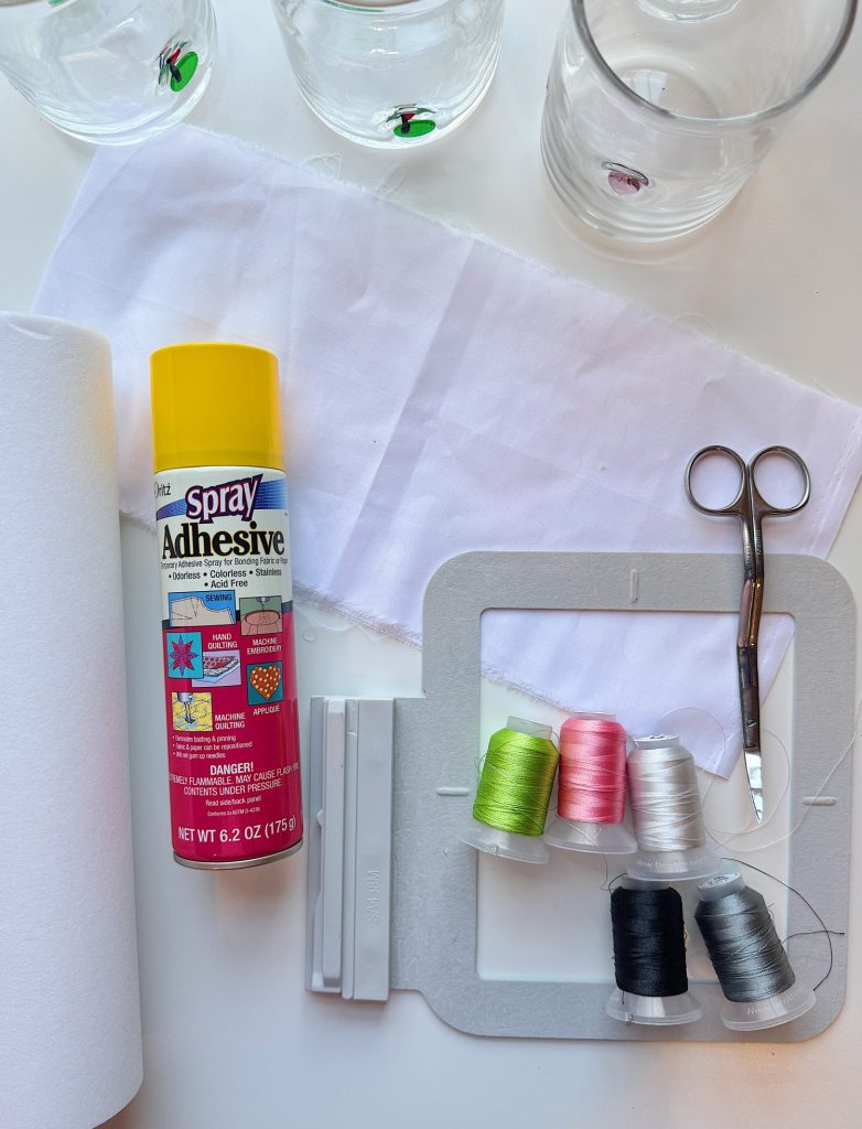 Sewing supplies on a table, including spray adhesive, fabric, scissors, embroidery frame, and spools of thread in various colors.