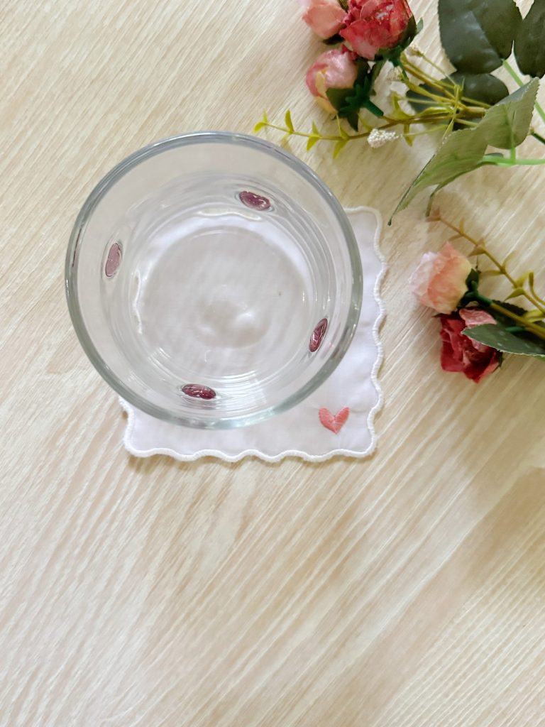 A glass of water on a square white coaster with a small pink heart, next to artificial pink and red flowers on a light wood surface.