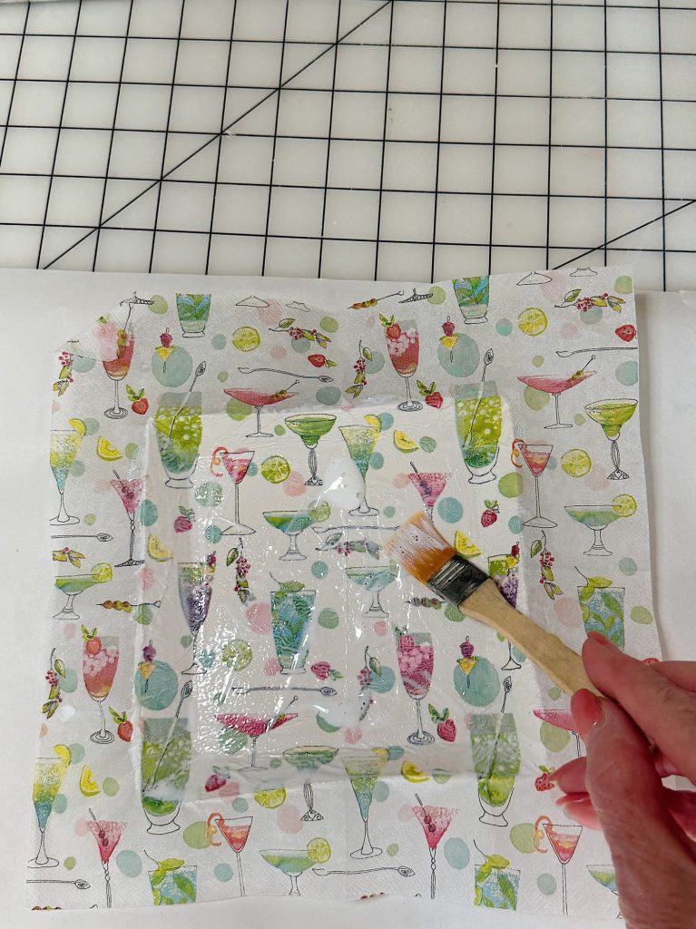 A hand uses a small brush to apply liquid onto a square piece of printed fabric with colorful cocktail designs, placed on a white surface and a grid background.
