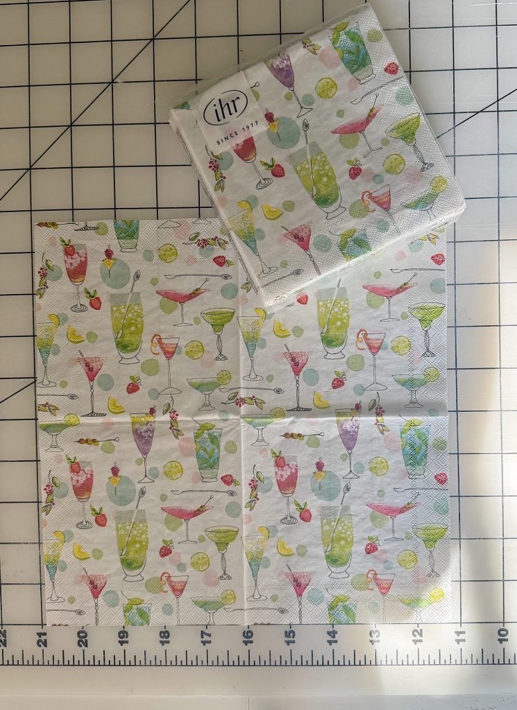 Four cocktail-themed paper napkins with colorful drink illustrations are laid out on a grid cutting mat, with a matching unopened napkin pack placed above them.