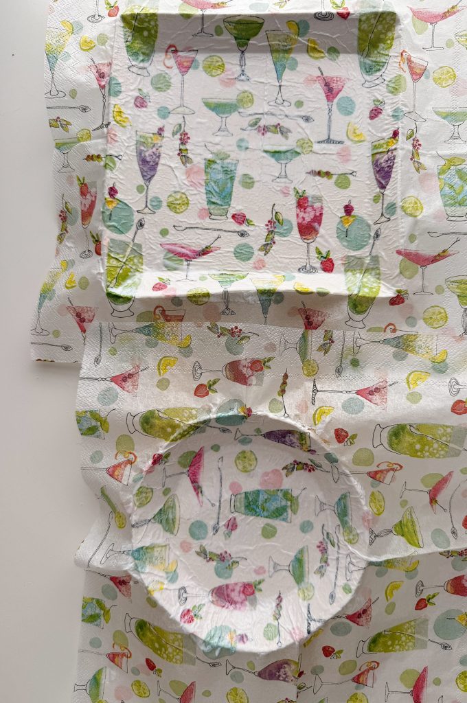 Square and round paper plates are placed on a patterned paper napkin featuring colorful cocktail glasses and lemon slices.