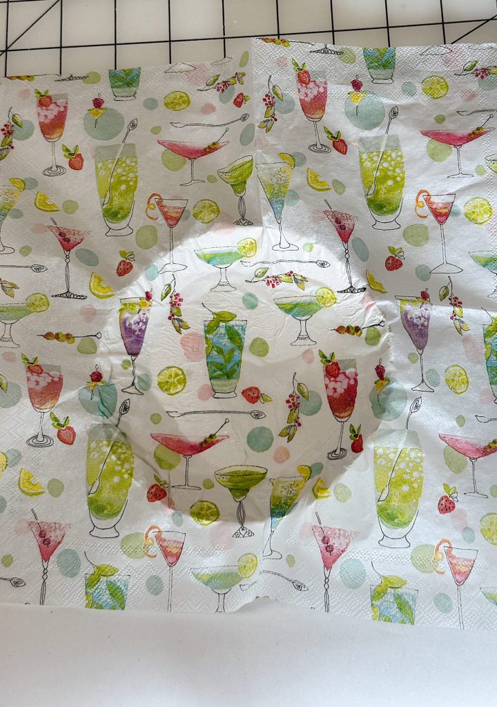 A white napkin decorated with colorful illustrations of various cocktails, fruits, and polka dots, shown on a checkered surface.