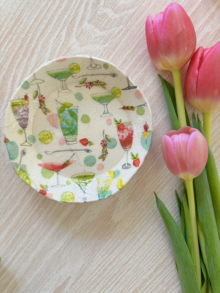 A small decorative plate with colorful cocktail glass designs next to three pink tulips on a light wooden surface.