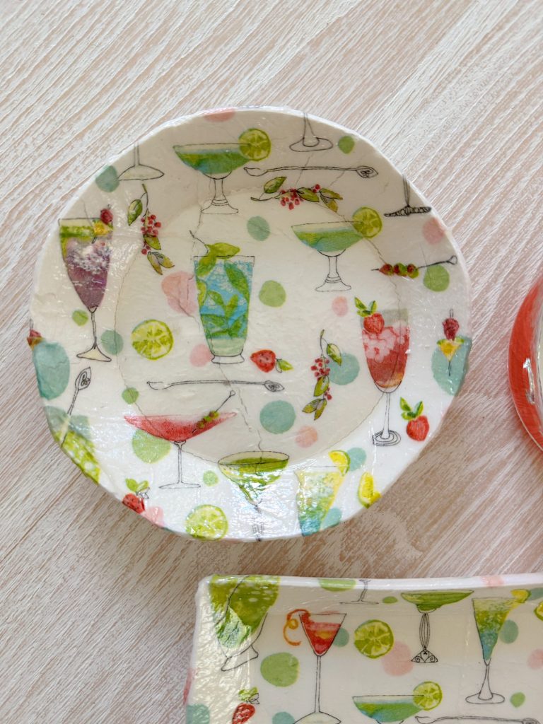 A small round plate decorated with colorful illustrations of various cocktails, fruit slices, and berries on a light background.