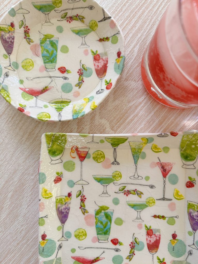 A round plate, a square plate, and a glass of pink beverage on a light wood surface; plates feature colorful cocktail and fruit designs on a polka dot background.