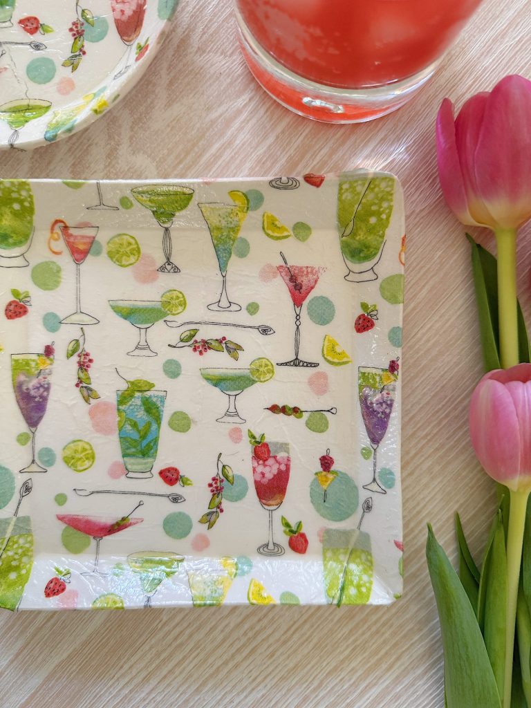 Square plate with colorful cocktail glass illustrations on a light table, next to a pink drink, a partial round plate, and pink tulips.