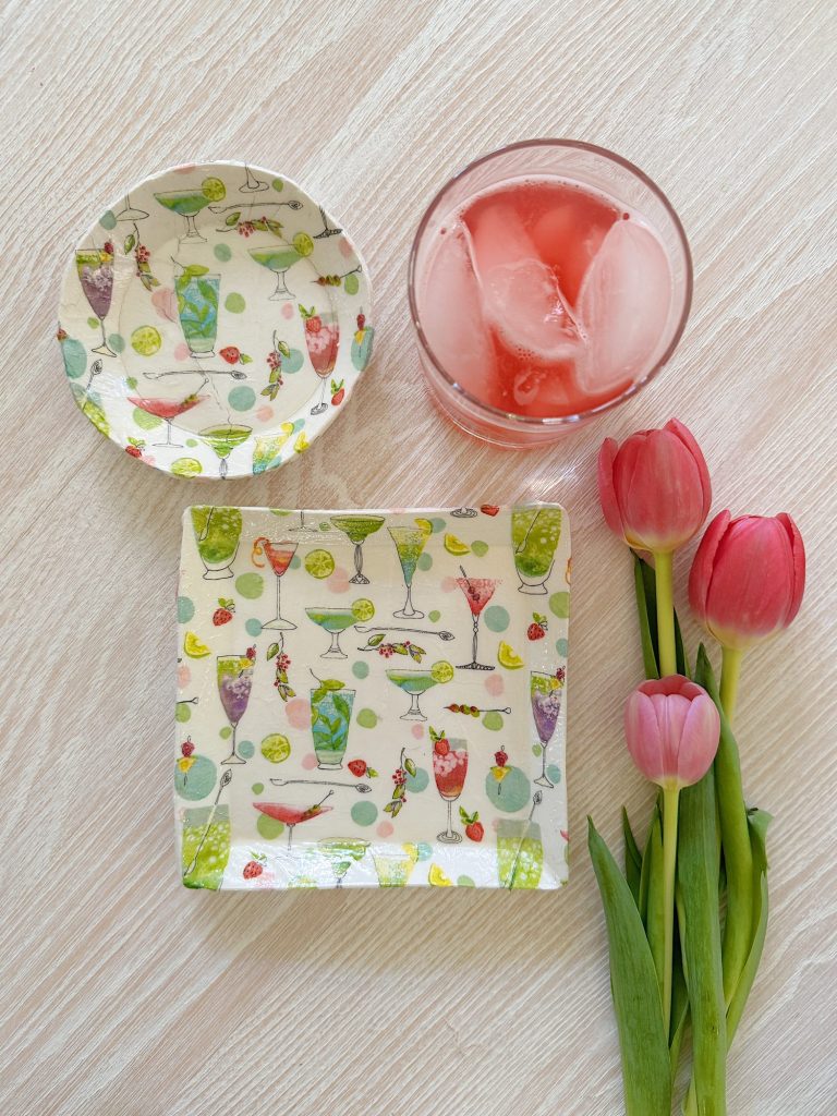 A glass of pink drink with ice, a round dish, a square plate with colorful cocktail designs, and three pink tulips on a light wooden surface.