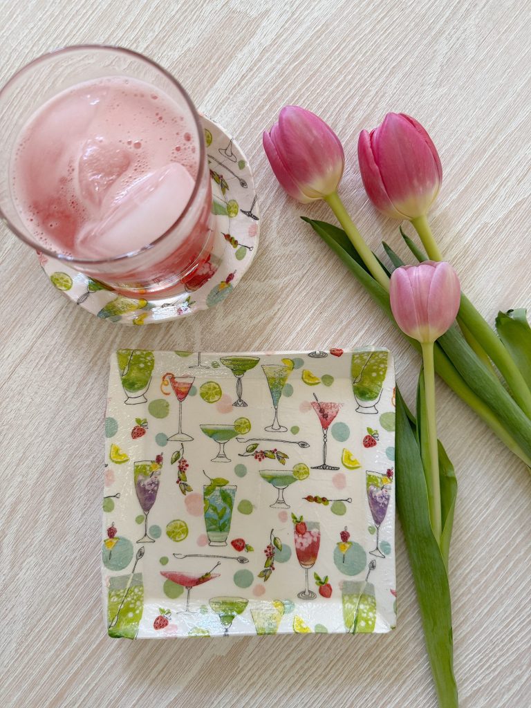 A glass of pink iced drink on a coaster, a square plate with cocktail illustrations, and three pink tulips on a light wooden surface.