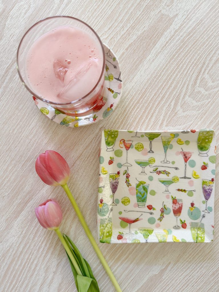 A glass of pink drink on a coaster, a matching plate with cocktail designs, and two pink tulips on a light wood surface.