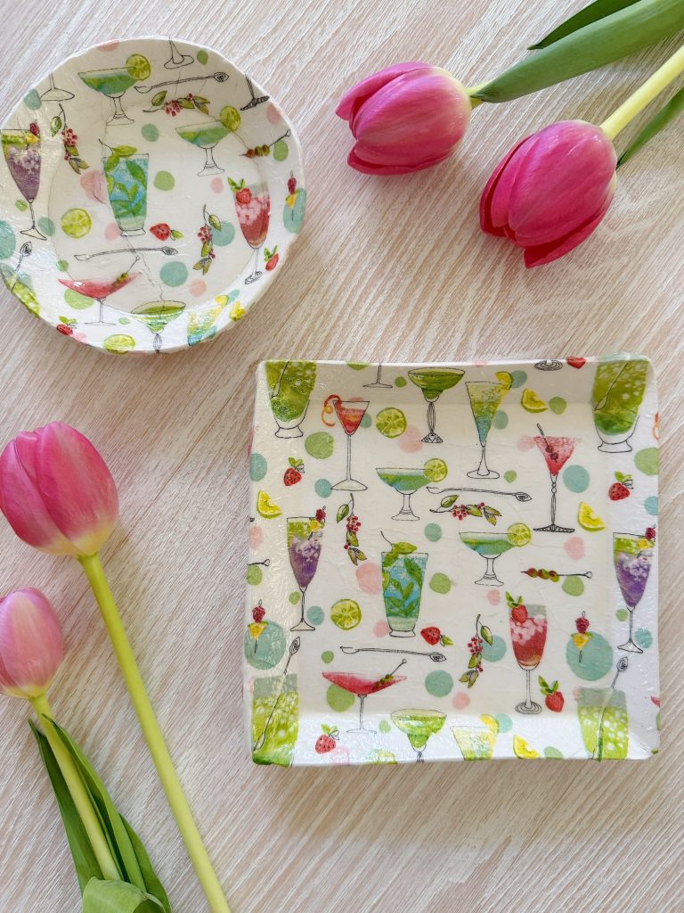 Two ceramic plates with colorful cocktail glass patterns are placed on a light wooden surface, surrounded by three pink tulips.