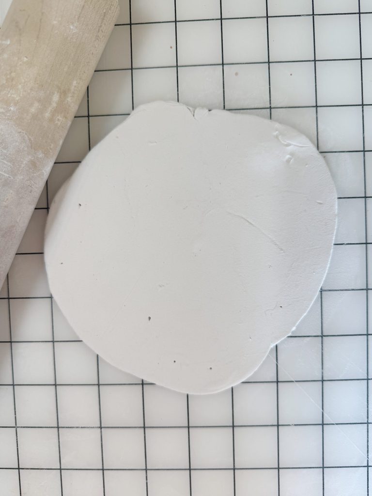 A sheet of white clay rolled flat on a grid-patterned surface with a wooden rolling pin visible on the left side.