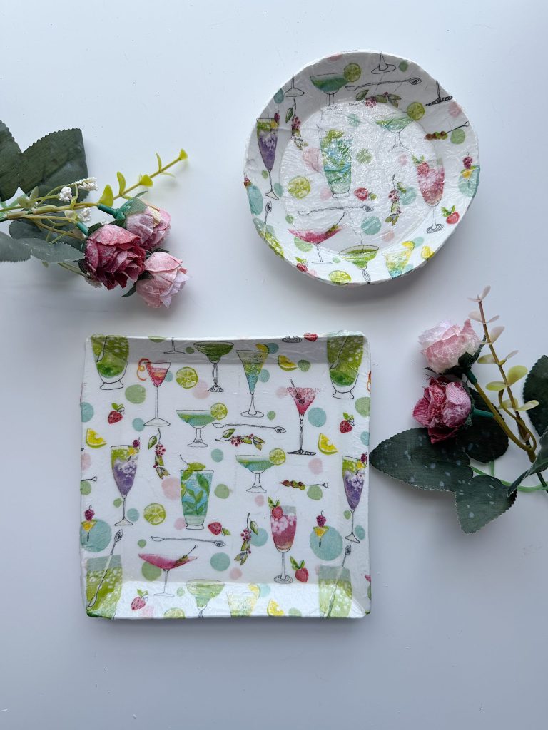 Two decorative plates, one round and one square, with colorful drink and fruit patterns, placed on a white surface alongside artificial pink flowers and green leaves.