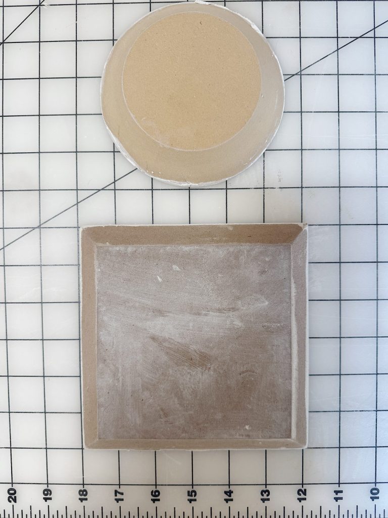 A round and a square piece of unglazed clay sit on a grid cutting mat, with measurement lines visible in the background.