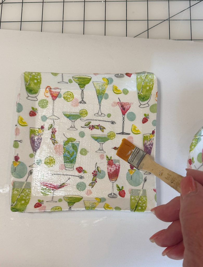 A hand uses a brush to apply a coat of glue or finish to a square plate decorated with colorful cocktail glasses and fruit patterns.