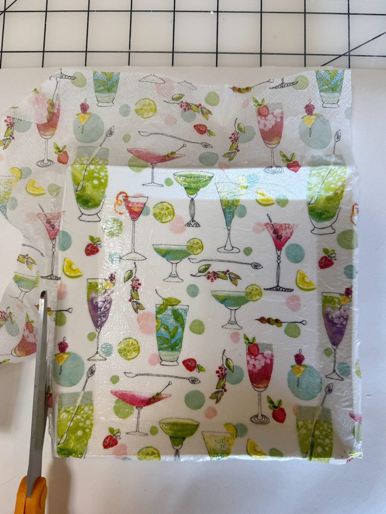 A hand uses scissors to cut a sheet of patterned wax wrap featuring colorful cocktail glasses, lemons, and cherries on a white surface.