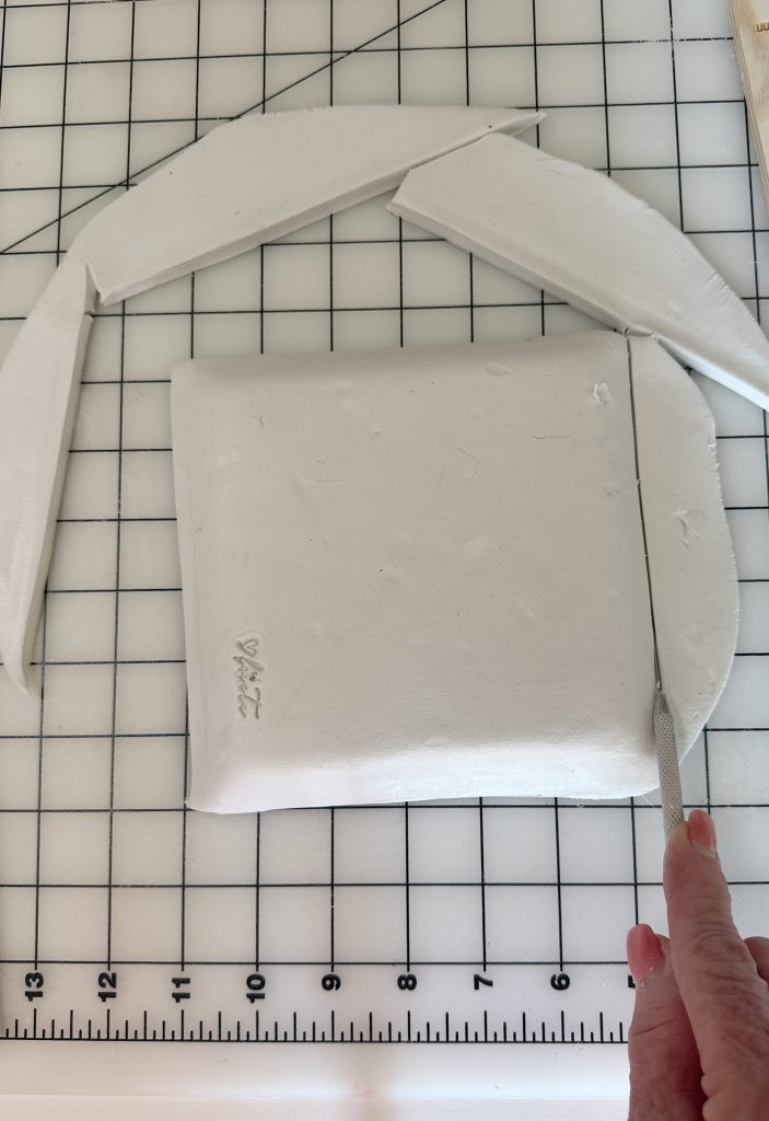A hand uses a craft knife to cut a rectangular piece of white clay, with additional clay pieces arranged above it on a grid cutting mat.